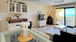 Una sala de estar con una mesa de centro de vidrio, un sofá, un televisor, un mueble con puertas de cristal y un balcón con sillas.
