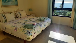 Un dormitorio con cama, almohadas, mesita de noche, lámpara, un cuadro y ventana con vistas.