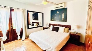 A hotel room with a bed, a ceiling fan, an air conditioning unit, a nightstand, a mirror, and a cityscape painting. - Villa Bali (Playa Blanca)