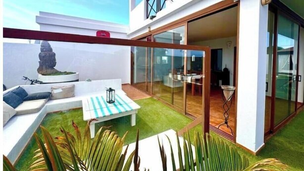 A modern outdoor living area with a glass door leading to a dining space, a small seating area with a table and chairs, and a decorative stone sculpture. - Villa Bali (Playa Blanca)