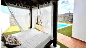 A bed with white bedding and a colorful pillow, a wooden ceiling, and a view of a swimming pool and garden. - Villa Bali (Playa Blanca)