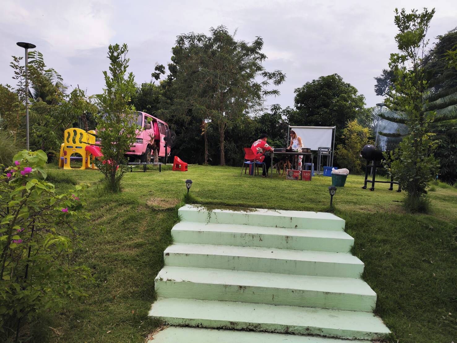 A grassy outdoor area with steps leading up to a white structure, surrounded by trees and a playground.