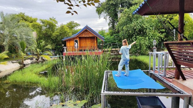 Un pontile in legno con una persona che pratica yoga, una casa in legno con tetto blu e uno stagno con ninfee.