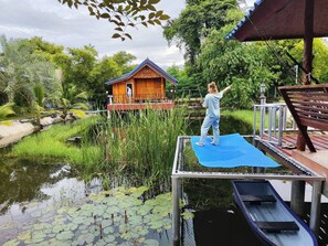 Eine Holzterrasse mit einer Person, die Yoga praktiziert, ein Haus mit blauem Dach und ein Teich voller Seerosen.