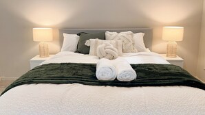 Ein ordentlich bezogenes Bett mit Kissen, Decke und aufgerollten Handtüchern. Auf beiden Seiten des Bettes stehen Nachttische mit Lampen.