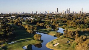 Luftaufnahme eines Golfplatzes mit mehreren Wasserhindernissen und Sandbunkern, umgeben von Bäumen und einer Stadtkulisse im Hintergrund.