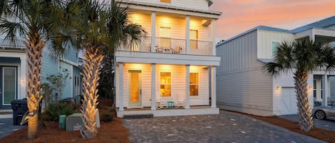 A two-story house with a balcony, surrounded by palm trees, and a paved driveway.