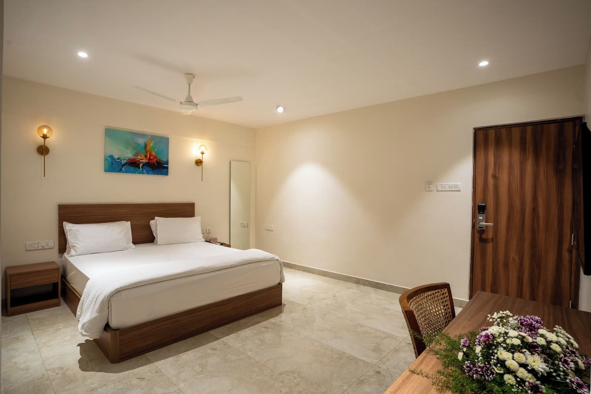 A bedroom with a bed, bedside tables, a ceiling fan, a wall-mounted light, a painting, and a dining table with flowers.