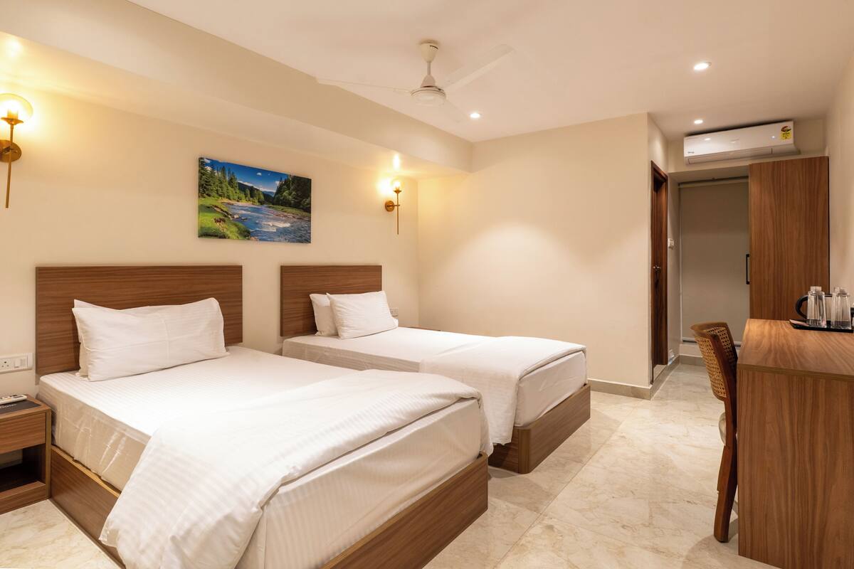 A hotel room with two beds, a desk, a chair, a painting, and an air conditioning unit.