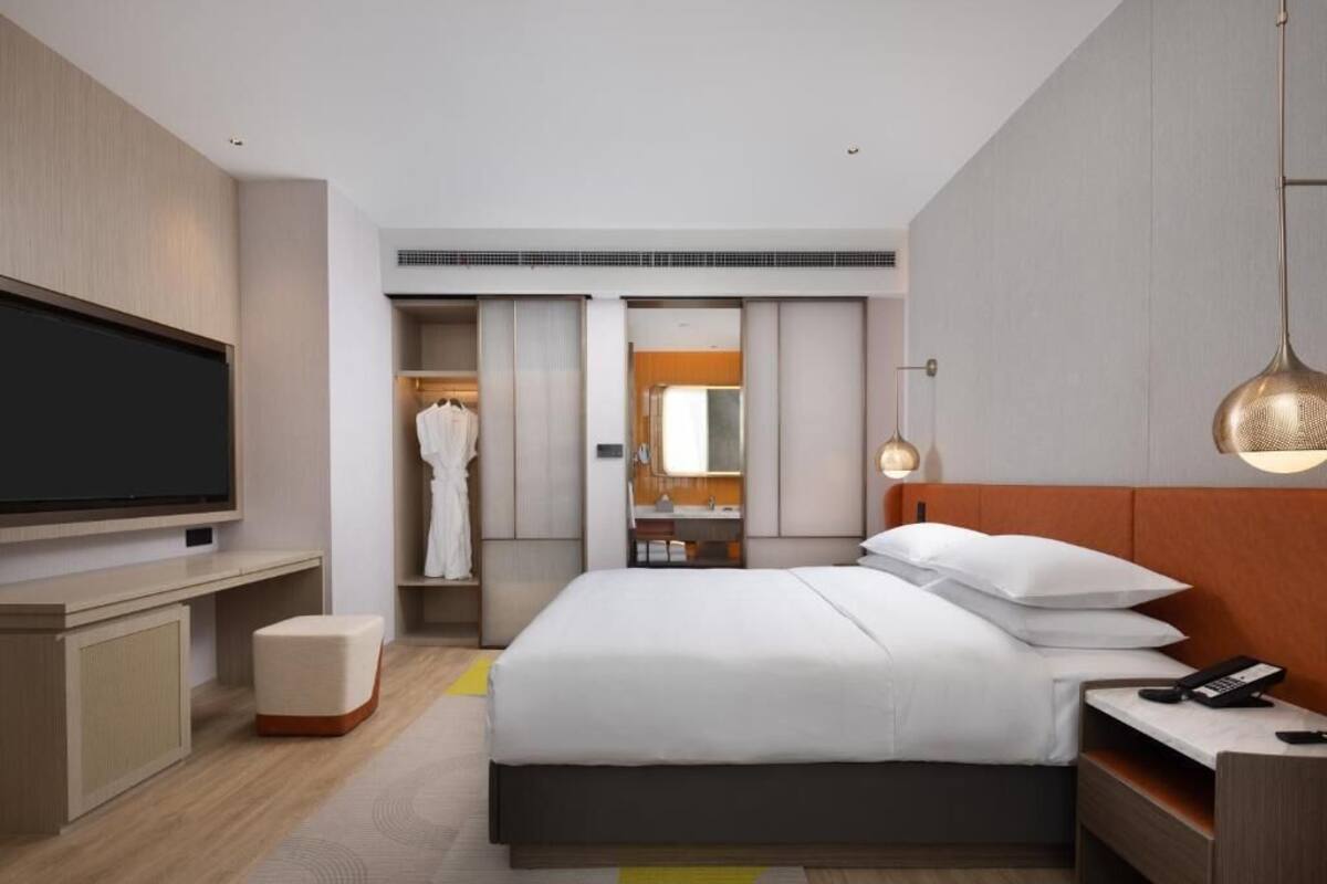 Una habitación de hotel moderna con un televisor de pantalla plana grande, una cama con ropa blanca, una mesita de noche con un teléfono y un armario con espejo.
