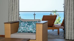 Un divano in vimini con un cuscino decorato, una sedia in legno e vista sull'oceano dal balcone.