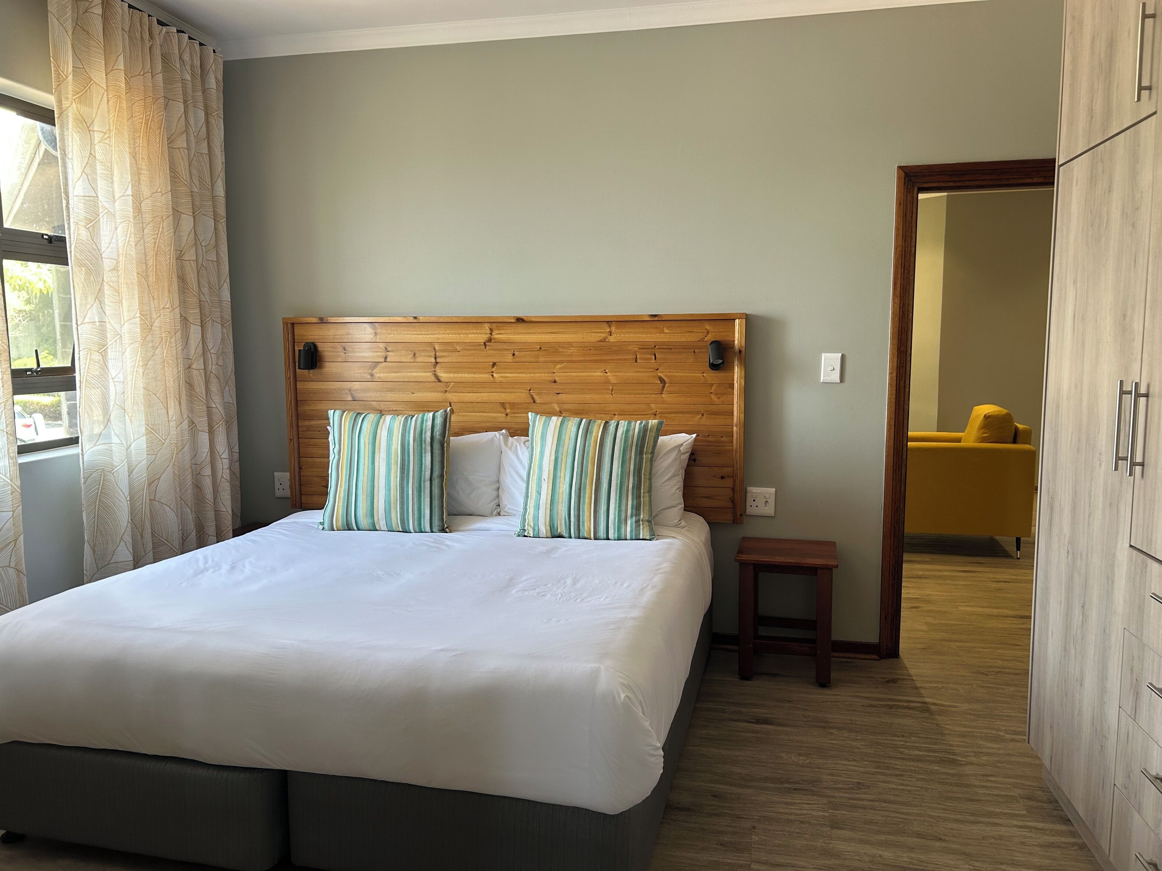 A bedroom with a wooden headboard, a bed with white bedding, striped pillows, a small wooden stool, and a wardrobe.