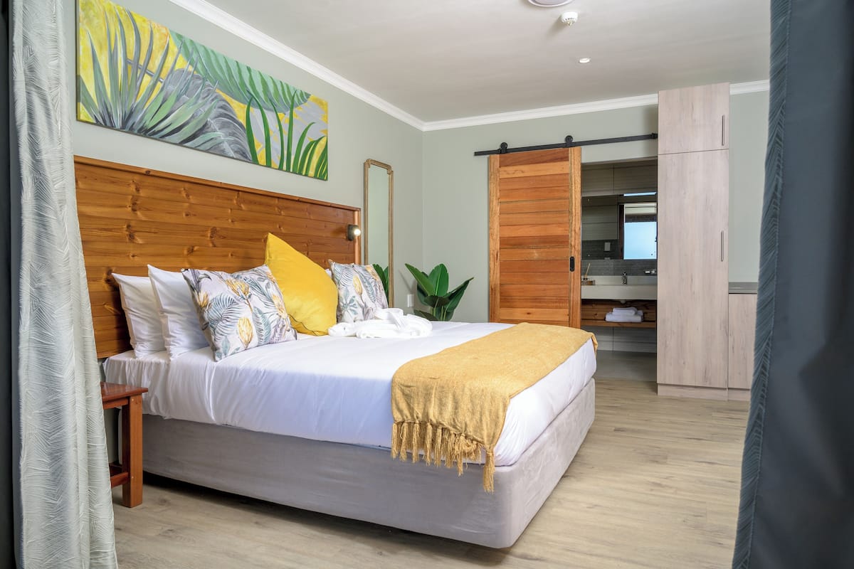 A bedroom with a large bed, a wooden headboard, a bedside table, a large mirror, a sliding door to a bathroom, and a painting on the wall.
