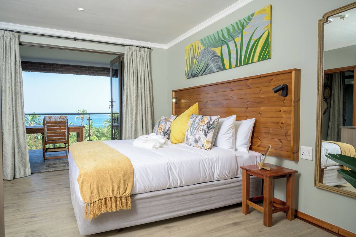 A bedroom with a large bed, a wooden headboard, a small table, a chair, a balcony with a table and chair, and a painting on the wall.