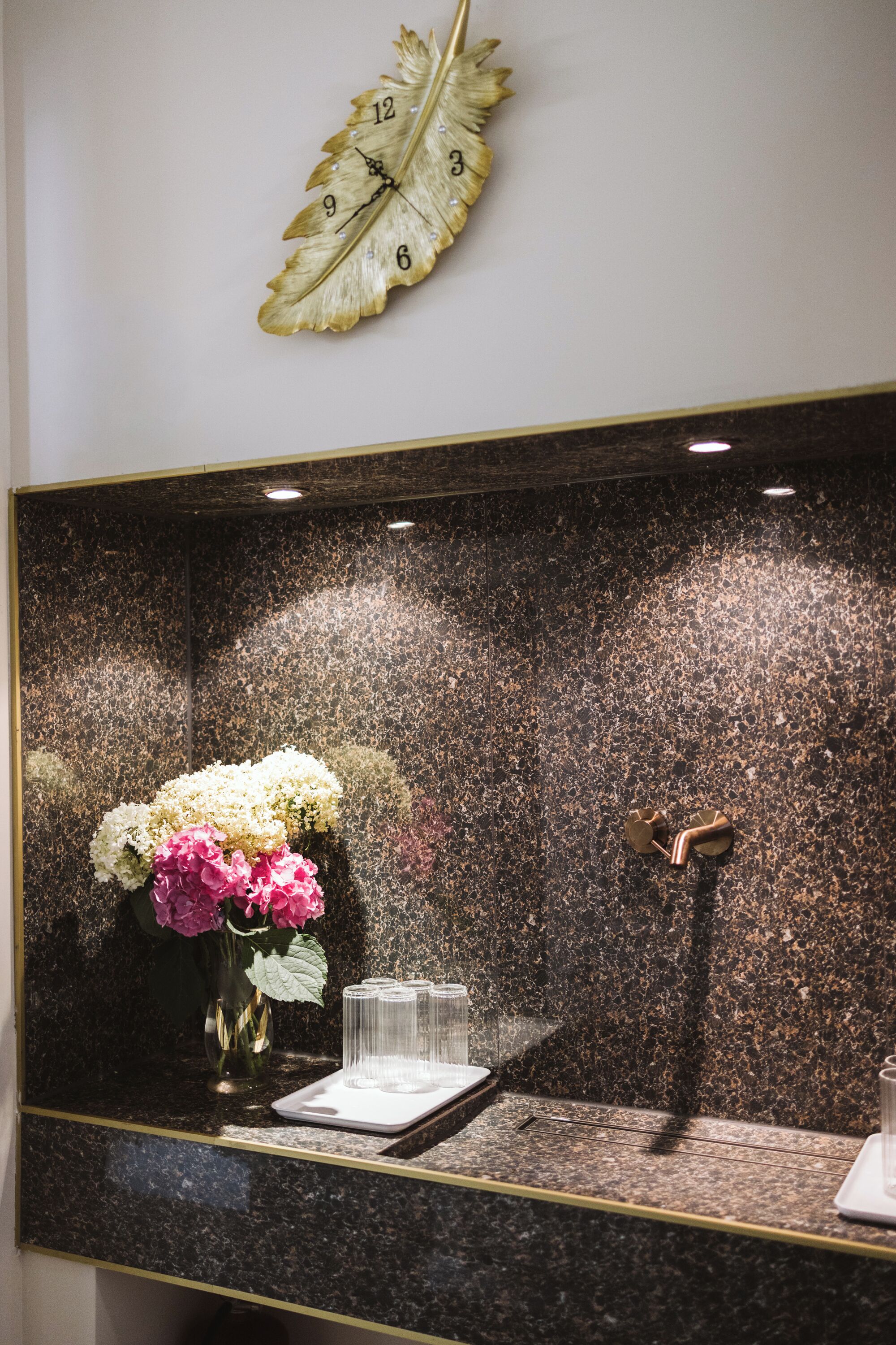 A wall-mounted clock with a leaf design, a mirror reflecting a vase with flowers, and a metallic door handle.