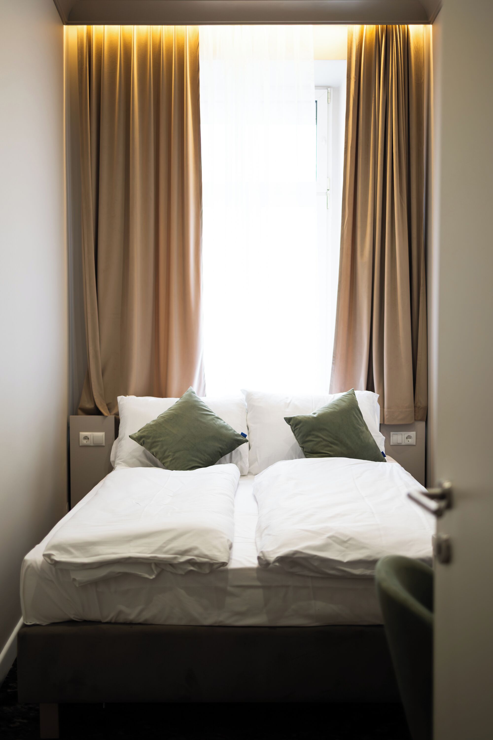 A bed with white linens and two pillows, a nightstand with a power outlet, and beige curtains.