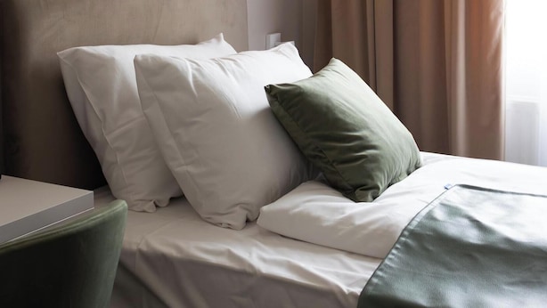 Een keurig opgemaakt bed met witte en groene kussens, een groene plaid en een hoofdbord met een verlicht paneel.