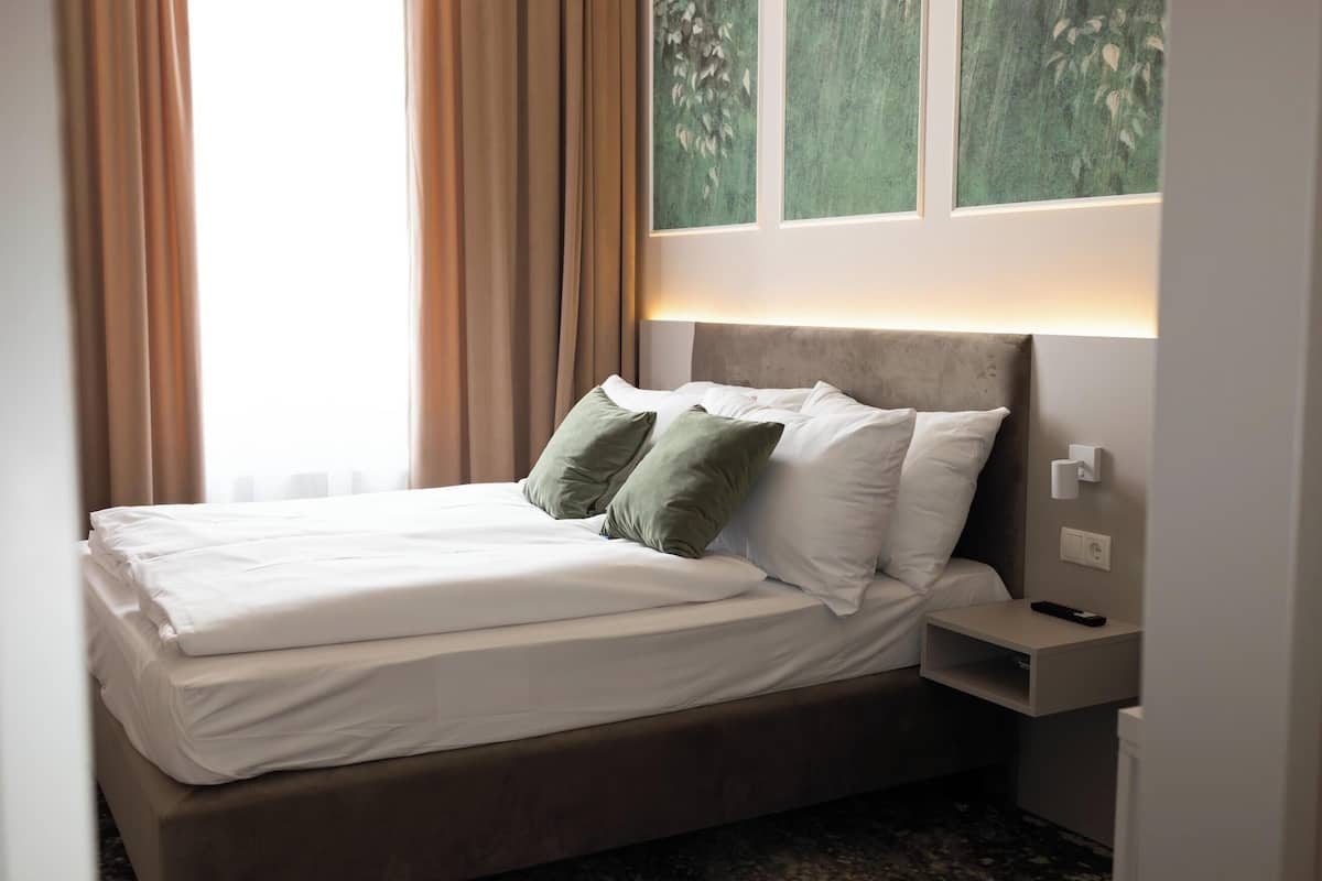 Un lit bien fait, avec du linge de lit blanc et des coussins verts, une table de chevet avec une télécommande et une fenêtre avec des rideaux.