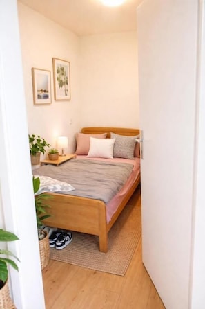 A bedroom with a wooden bed, a bedside table with a lamp, a wall-mounted shelf, and a door leading to another room. - Zurich Airport Cozy Studio (Oberglatt)
