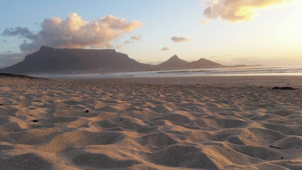 Ein sandiger Strand mit wellenartigen Mustern, ein ruhiges Meer und ferne Berge unter einem teils bewölkten Himmel.