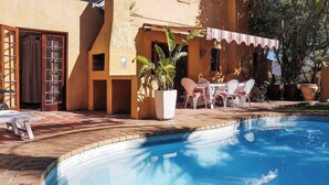 Ein Pool mit gefliestem Rand, eine Terrassenfläche mit Stühlen und Tisch sowie ein Gebäude mit Tür und Fenstern.
