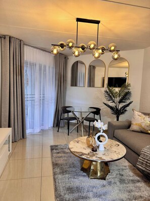 Ein modernes Wohnzimmer mit rundem Couchtisch, einer Couch, einem Essbereich mit Stühlen, einem großen Spiegel und einer Deckenleuchte.