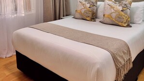 Una cama bien tendida con una paleta de colores blanco y beige, un colgante de luz y una ventana con cortinas translúcidas.