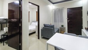 A hotel room with a bed, a desk, a chair, a small table, a potted plant, and a window with curtains. - Manzil Anilao Unit 1 - Ground Floor 1st Room (Mabini)