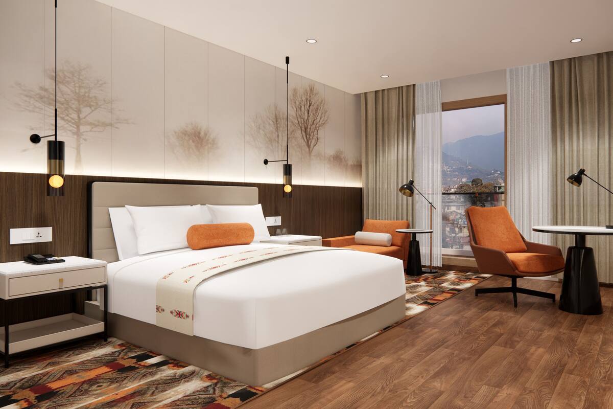 Una moderna camera d'albergo con un letto grande, un'area salotto con una poltrona e un tavolino, un comodino e vista sulla città attraverso una finestra.