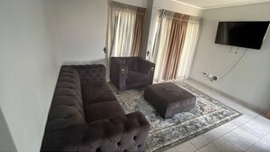 Uma sala de estar com sofá, poltrona, banquinho, tapete estampado, cortinas e televisão fixada na parede.