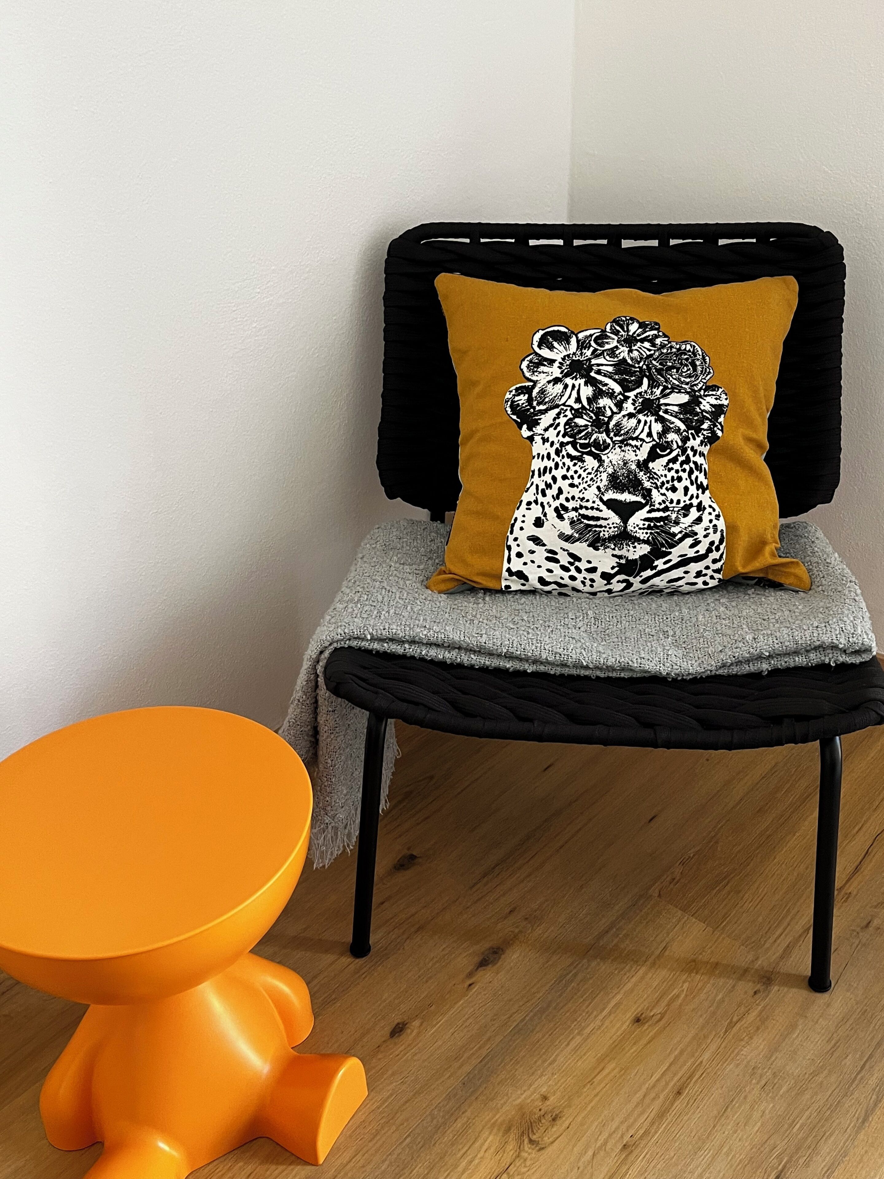 Een stoel met een zwart frame en een mosterdkleurig kussen, een klein oranje krukje en een grijze gebreide plaid.