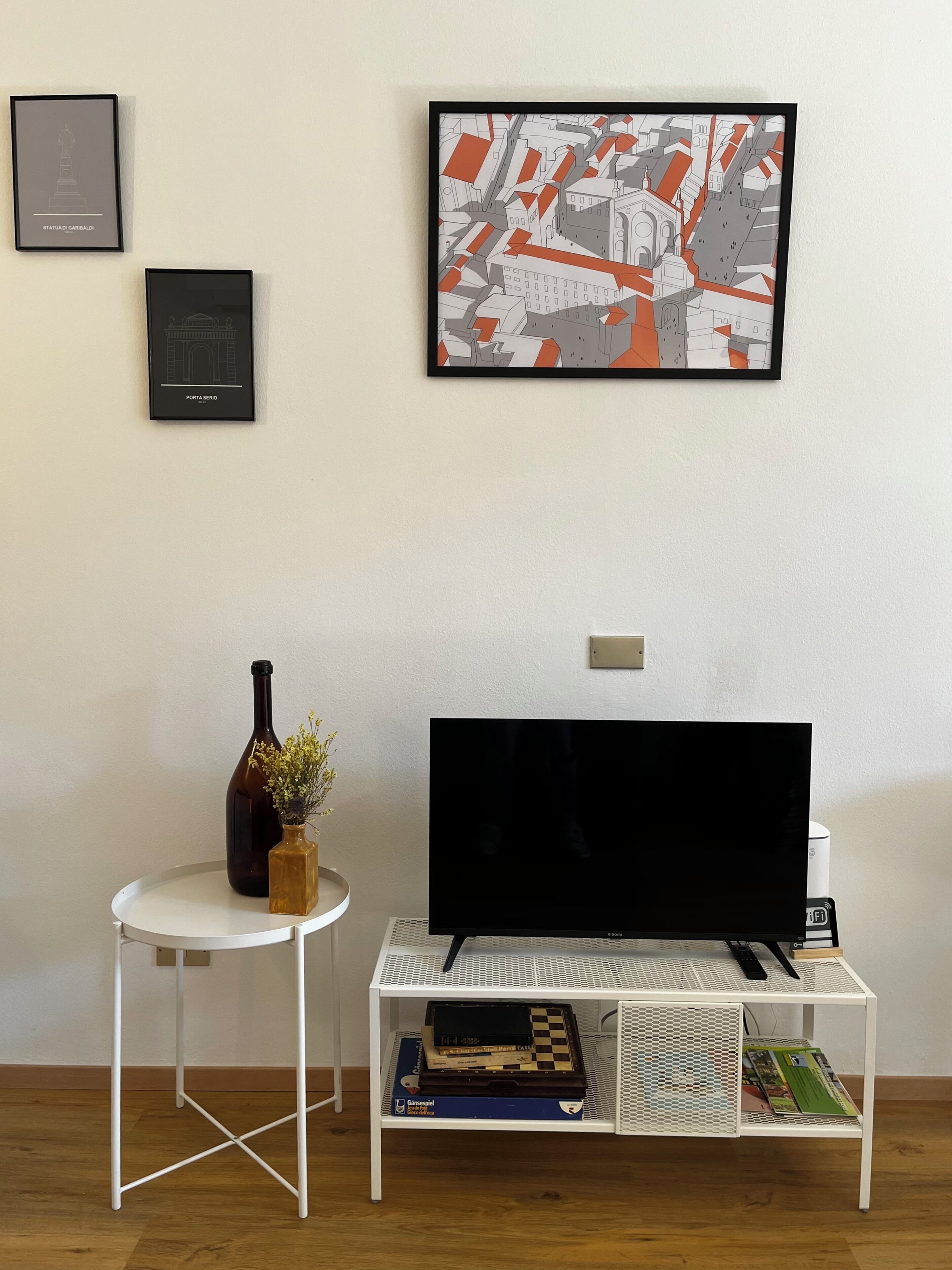 Una stanza con una televisione su un mobile bianco, un tavolino con un vaso e libri, e due quadri appesi al muro.