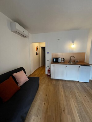 A compact living space with a kitchenette, a black sofa with cushions, and a wooden floor.