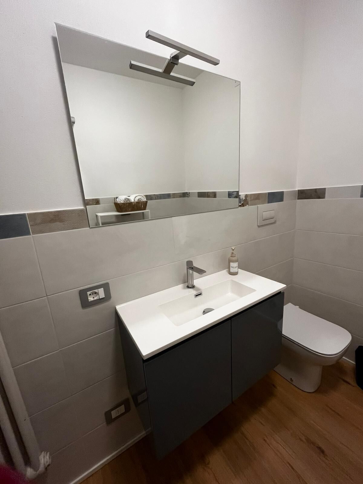Une salle de bain avec un lavabo blanc, des meubles sombres, des toilettes et un miroir surmonté d’un luminaire.