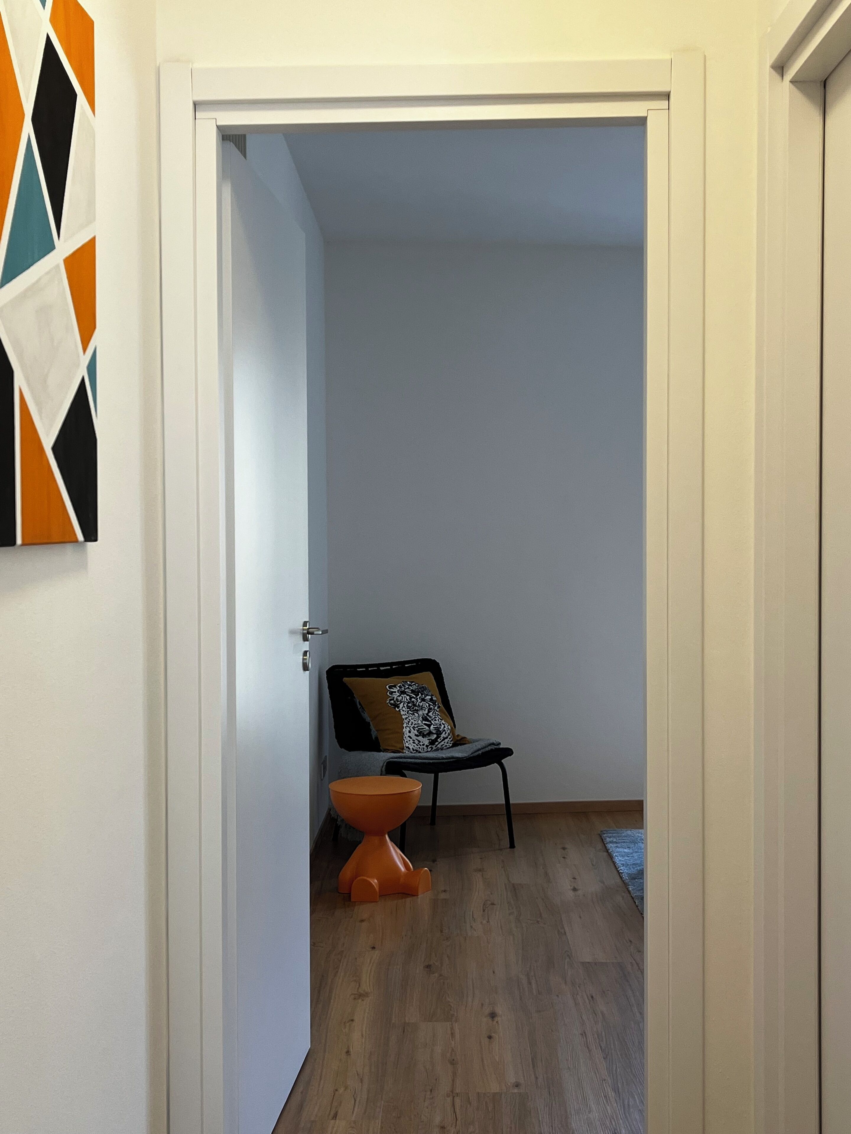 Un pasillo con una puerta que da a una habitación, una silla con cojín y un taburete pequeño naranja.