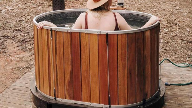 Una persona relajándose en un jacuzzi de madera al aire libre, rodeada de árboles.