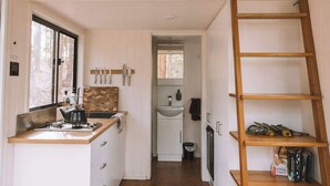 Cocina compacta con estufa a gas, un lavamanos y una ventana. Una escalera de madera está a la derecha, y una estantería a la izquierda.