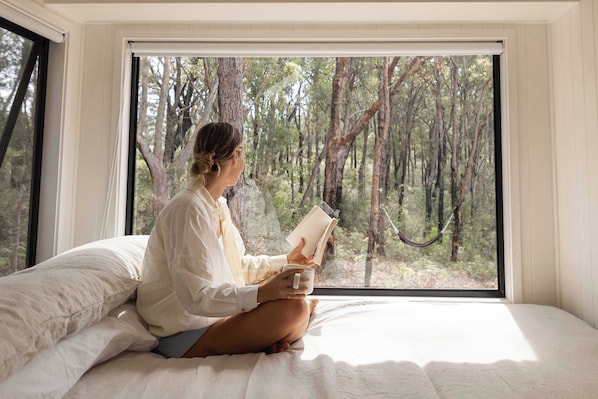 A person sitting on a bed in a room with a large window overlooking a forest.