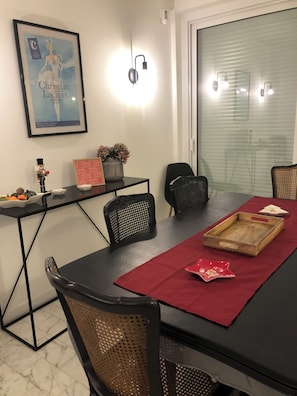 Uma sala de jantar com mesa preta, jogo americano vermelho e cadeiras pretas. Há um quadro na parede, uma pequena mesa com um vaso e um soldadinho de chumbo, e uma janela com persianas.