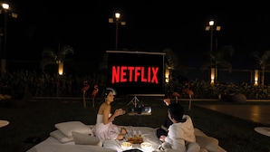 Escena nocturna al aire libre con una pantalla grande mostrando 'NETFLIX' y dos personas sentadas en un sofá mirando la pantalla.