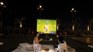 Un área de estar al aire libre de noche con una gran pantalla mostrando un partido de cricket, rodeada de palmeras y flamencos.