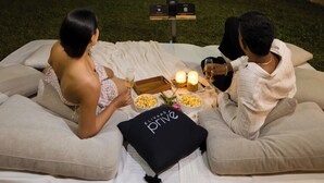 Una pareja descansando en una cama con una pantalla grande mostrando 'NETFLIX' al fondo.
