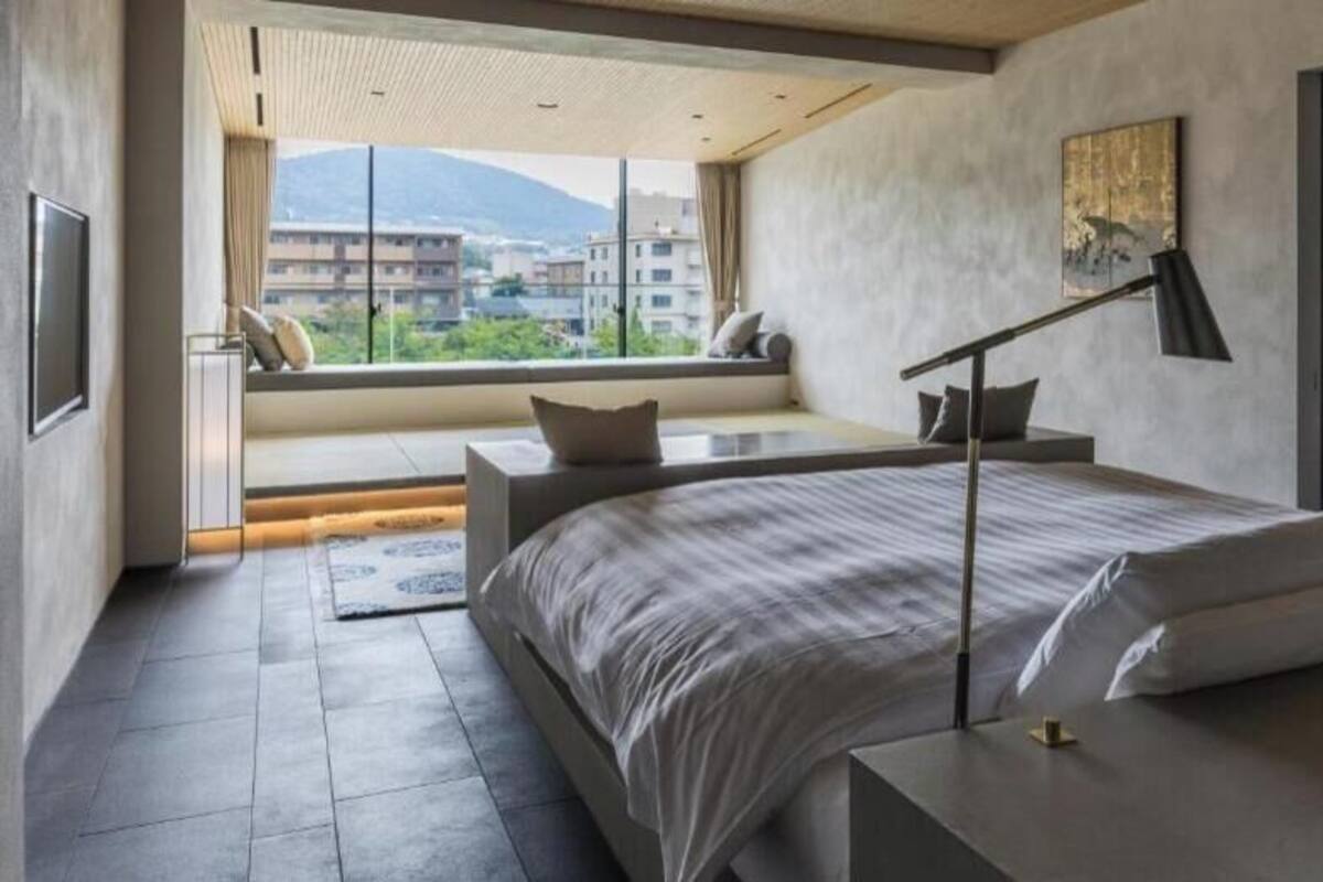 A modern bedroom with a large bed, a bedside table, a nightstand, a window with a view of buildings, and a wall-mounted shelf.