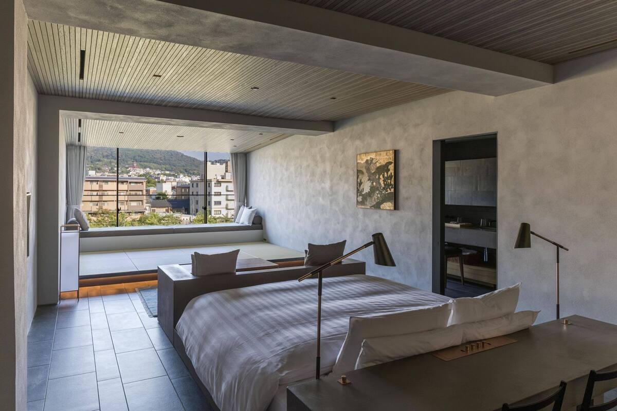 A modern bedroom with a large bed, a desk, a chair, a sofa, and a view of a cityscape through a large window.