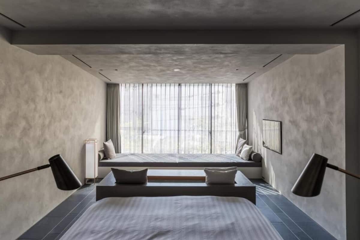A modern bedroom with a large bed, a bench, and a window with sheer curtains.