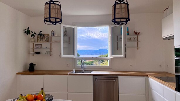 Une cuisine avec vue, des meubles blancs, un lave-vaisselle en acier inoxydable et un panier de fruits posé sur le plan de travail.