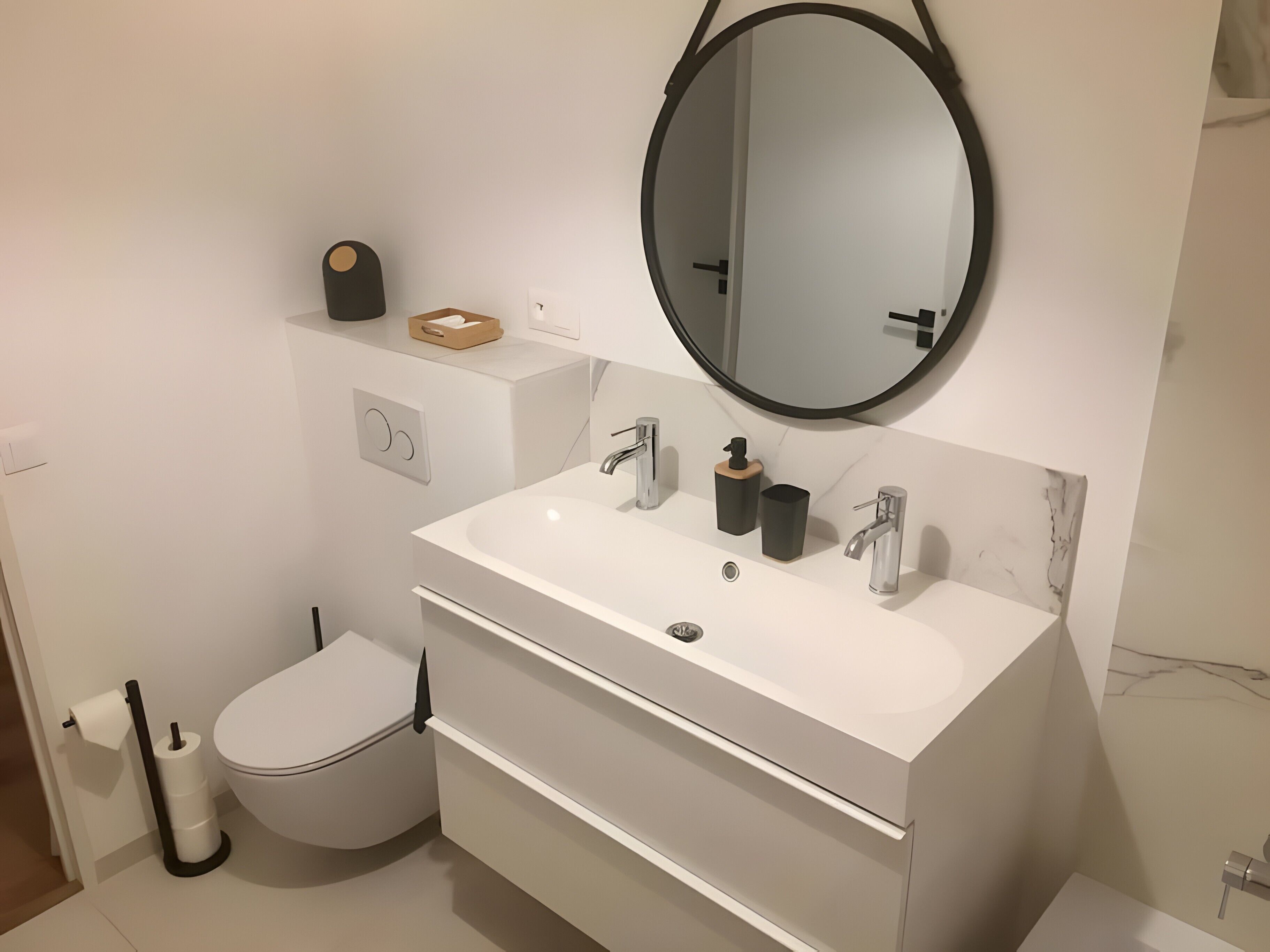 Une salle de bain moderne avec un lavabo blanc, un miroir rond, des toilettes et un bidet.