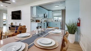 Spiseplads med et bord dækket til fire personer, et køkken i baggrunden og en stue med sofa og fjernsyn.
