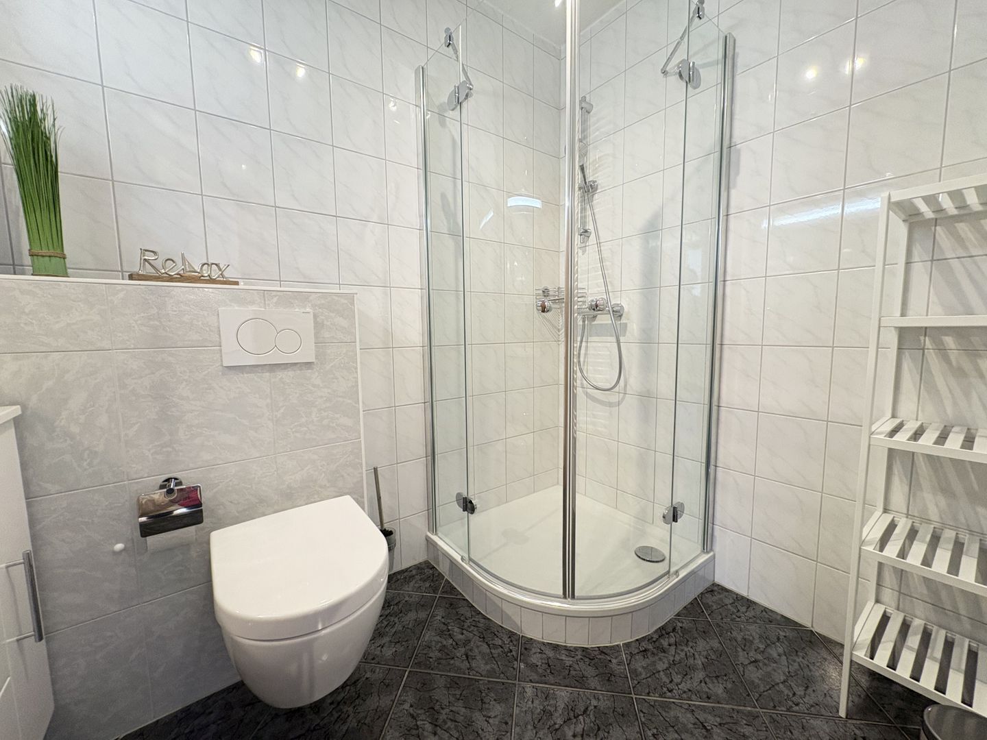 A modern bathroom with a glass-enclosed shower, a toilet, and a white shelf unit.