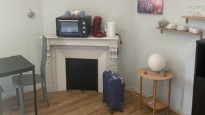 Habitación compacta con chimenea blanca, microondas, tetera roja, una mesita con una maleta azul y estante en la pared con cristalería.